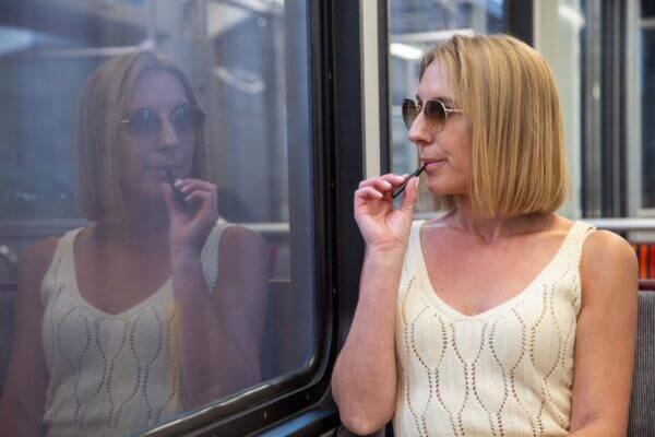 Woman in sunglasses and white top vaping on a train, reflected in window.