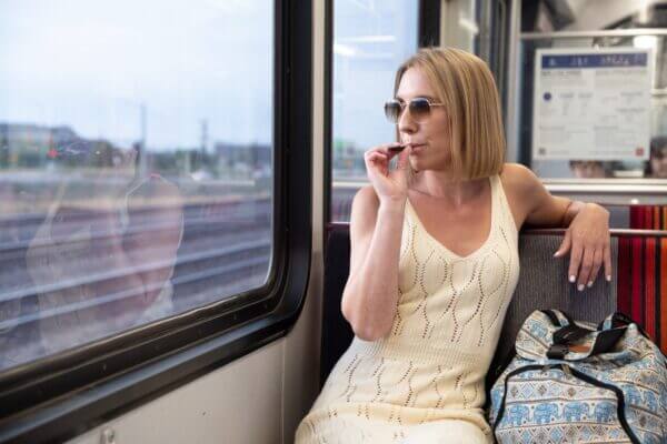 Woman in sunglasses vaping on a train, wearing a white dress, with a patterned backpack, looking out the window.
